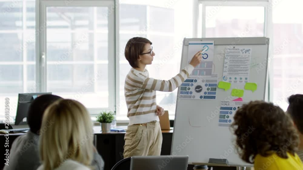 Tilt down of young caucasian businesswoman in eyeglasses standing by flipchart in the office and giving a presentation to multiethnic employees during corporate education Tilt down of young caucasian businesswoman in eyeglasses standing by flipchart in the office and giving a presentation to multiethnic employees during corporate education