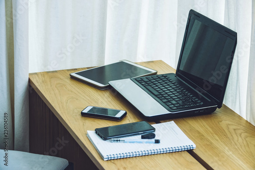 Interior detail view of a laptop computer on a home living room coffee table, with a telephone and tablet near a window with curtains.