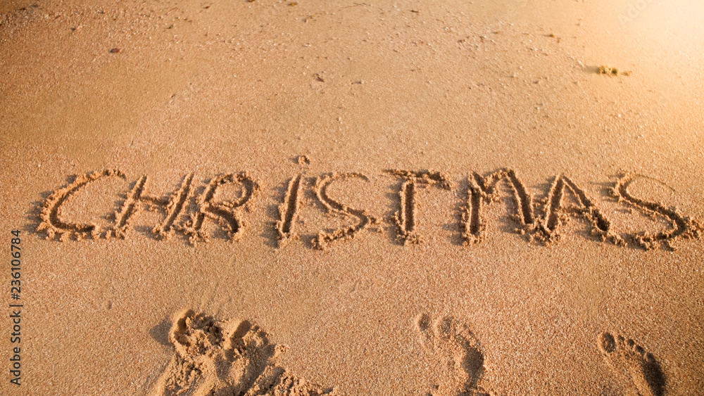 Fototapeta premium Photo of word Christmas written on sand beach at ocean. Concept of winter holidays, New Year and tourism