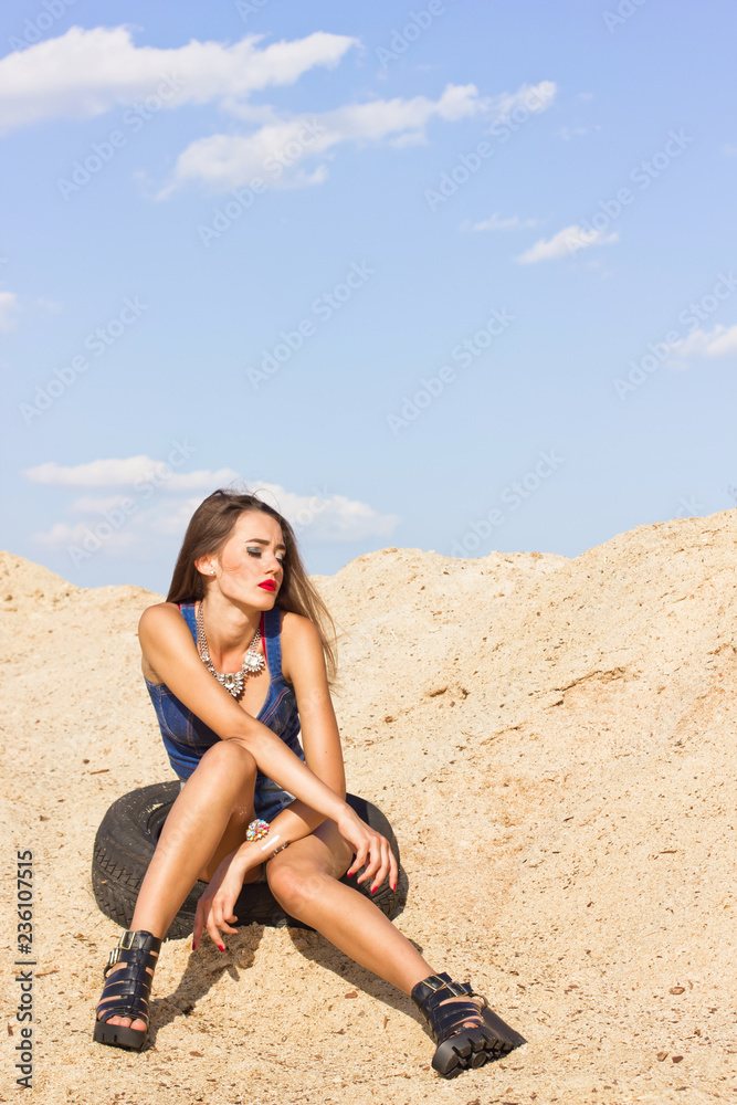 Summer heat. A beautiful young tanned girl in denim clothes and jewelry is sitting on a car tire, against the background of sawdust and sky