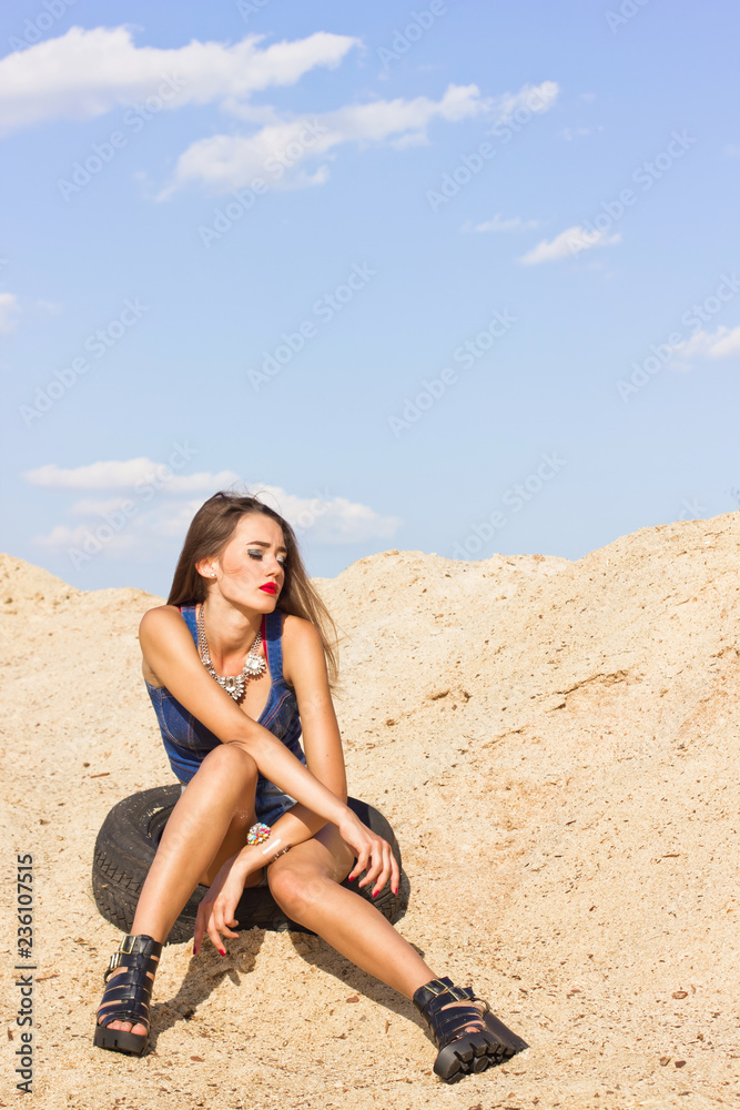 Summer heat. A beautiful young tanned girl in denim clothes and jewelry is sitting on a car tire, against the background of sawdust and sky Summer heat. A beautiful young tanned girl in denim clothes and jewelry is sitting on a car tire, against the background of sawdust and sky