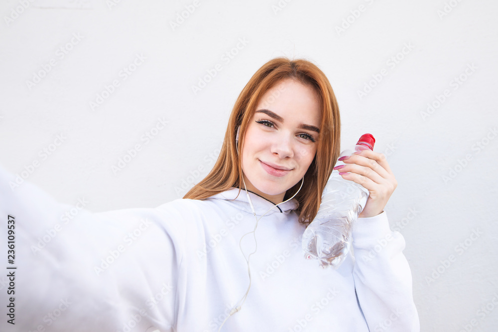 Portrait of an attractive young girl holding a bottle of water and makes selfie against the background of a white wall. Sports concept. Morning run Portrait of an attractive young girl holding a bottle of water and makes selfie against the background of a white wall. Sports concept. Morning run