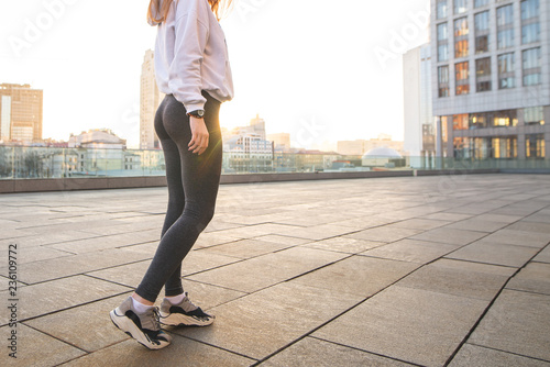 Photo of a girl's legs in sportswear walking around the city. Close-up photo of the bottom of a sports girl in leggings against the background of a city landscape at sunset.