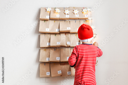 Little baby Santa's helper is watching the advent calendar for Christmas. Mood Board with gifts and star garland .