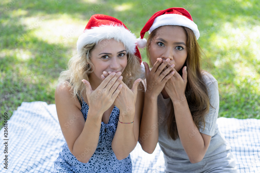 Surprised women wearing Santa hats and covering mouths in park. Playful pretty ladies looking at camera and sitting on grass. Women friendship and Christmas concept. Front view. Surprised women wearing Santa hats and covering mouths in park. Playful pretty ladies looking at camera and sitting on grass. Women friendship and Christmas concept. Front view.
