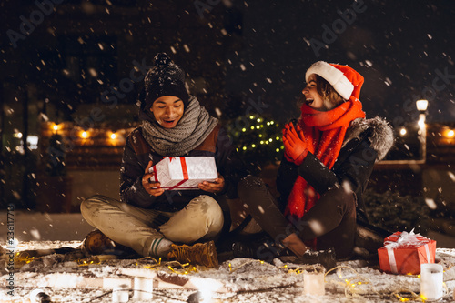 Emotional excited young loving couple sitting outdoors in evening in christmas hat holding gift box.