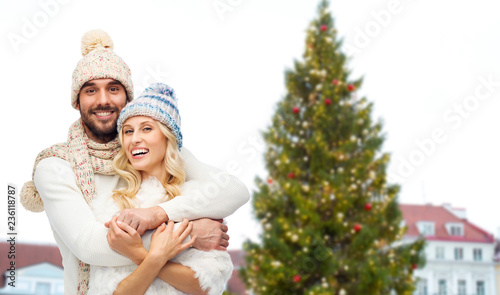 winter and holidays concept - happy couple in hats hugging over christmas tree at tallinn old town hall square background