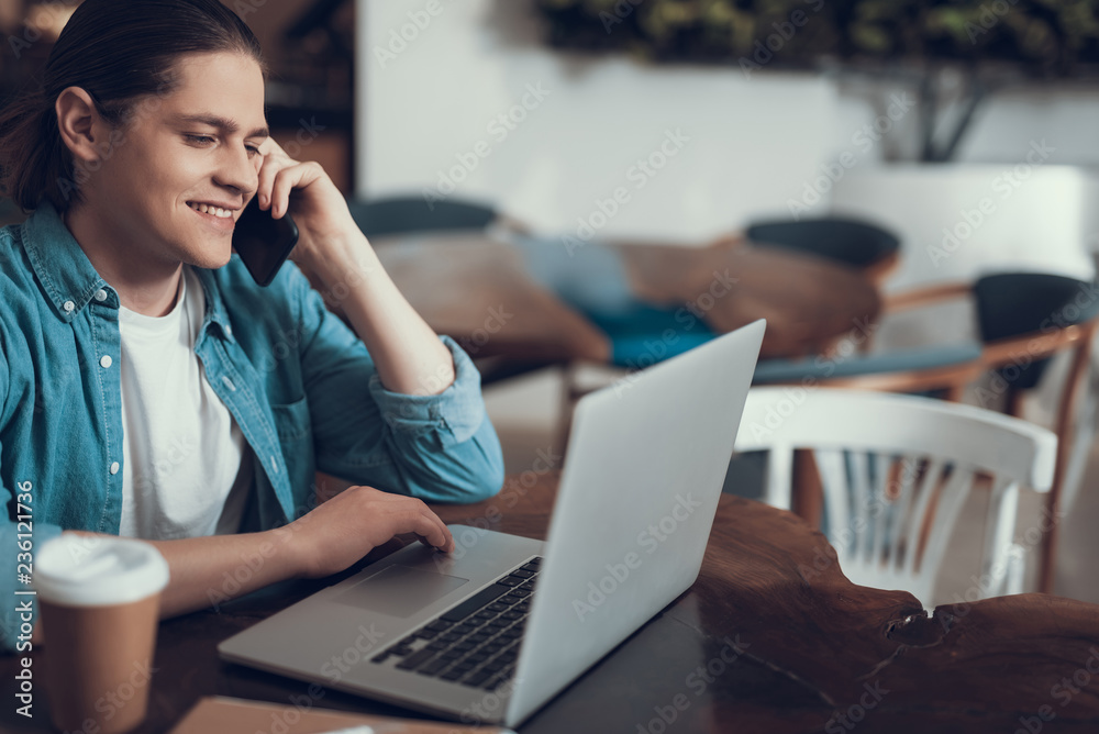 Cheerful young man talking on the phone while using modern laptop Cheerful young man talking on the phone while using modern laptop