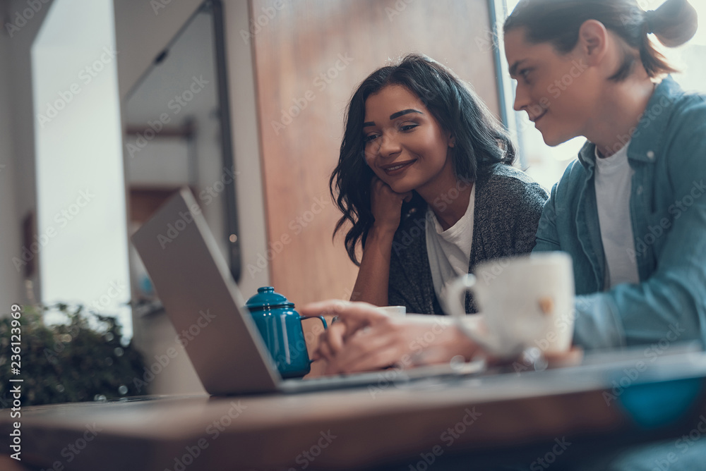 Cheerful young woman sitting near her smiling boyfriend and feeling interested while looking at the screen of a modern laptop Cheerful young woman sitting near her smiling boyfriend and feeling interested while looking at the screen of a modern laptop