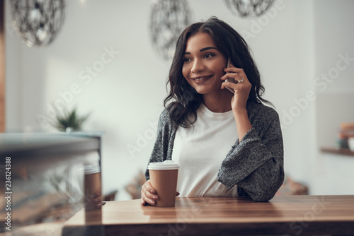 Peaceful relaxed young lady smiling and holding her carton cup of coffee while having a pleasant phone talk in a cafe