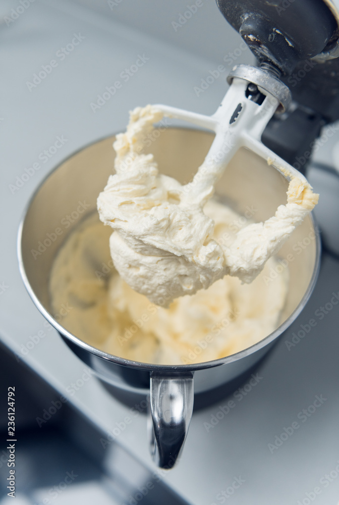 An industrial kitchen food mixing machine whipping and mixing sweet creamy fluffy light cake mixture, in a silver metal bowl.