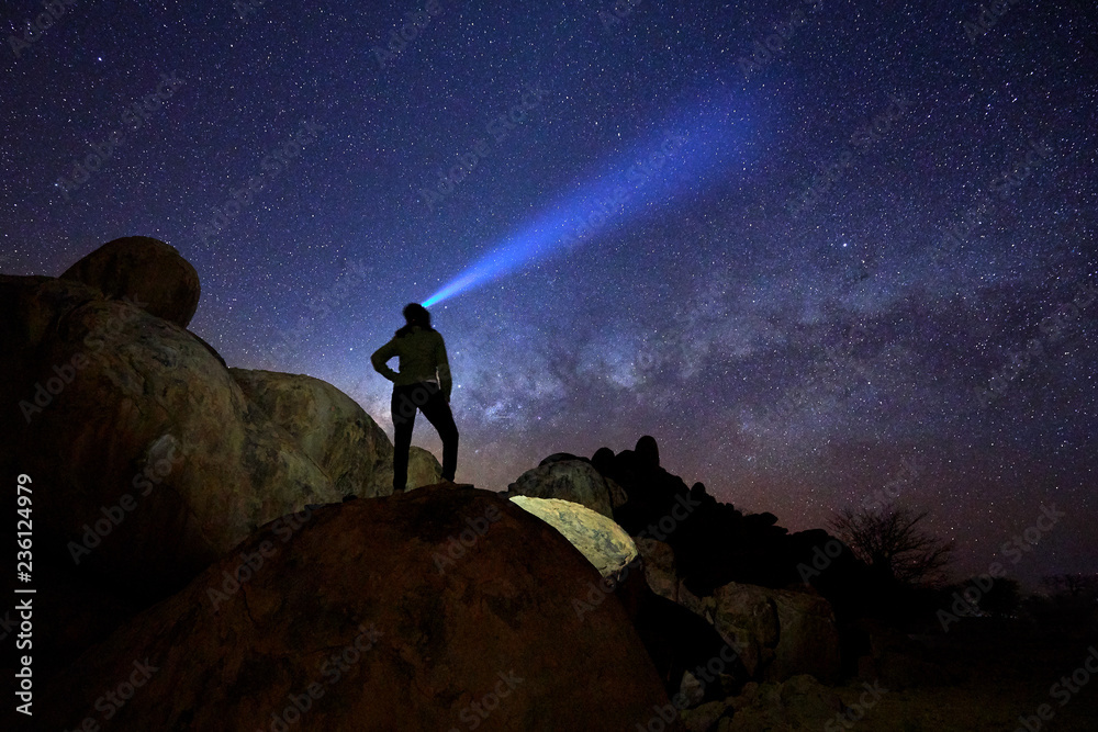 Silhouette of lone woman on rounded rocky structures gazing at milky way and the stars,  focused beam of headlamp pointing directly to milky way. Night sky photo. Twyfelfontein, Damaraland, Namibia.