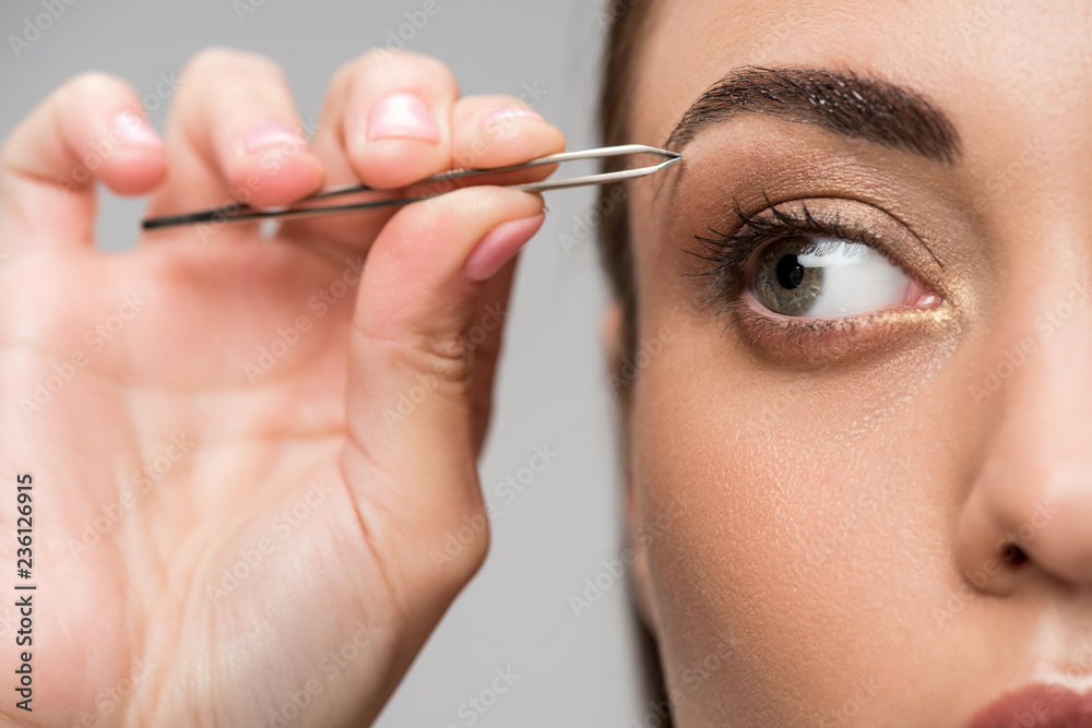 close up of young woman correcting shape of eyebrows with tweezers isolated on grey
