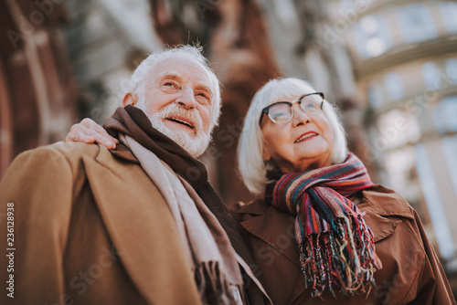 Low angle portrait of senior lady in glasses hugging husband. They looking away and smiling