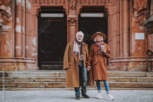 Full length portrait of stylish bearded man and his wife standing near old building. Lady in hat holding cup of coffee