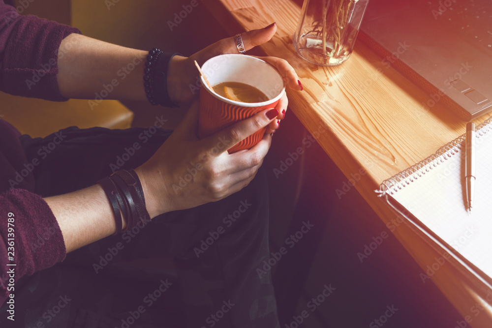 Top view. Student woman holds with two hands a hot cup of tea with Hippophae and a slice of orange. Workplace, notebook with pen and laptop, the working process. coffee shop, warm color toning