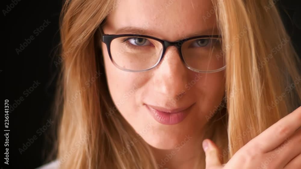 Close-up shy face of blonde caucasian spectacled girl, looking natural at camera in motion and cute covering herself with hair, black background Close-up shy face of blonde caucasian spectacled girl, looking natural at camera in motion and cute covering herself with hair, black background