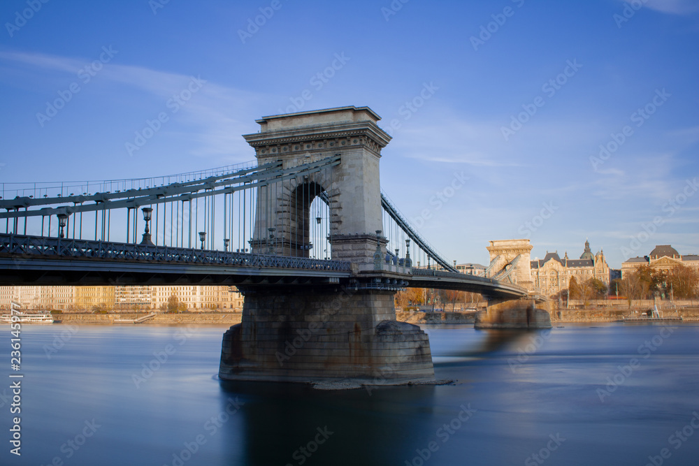 Obraz premium Die Kettenbrücke über die Donau in Budapest, der Hauptstadt von Ungarn am späten Nachmittag kurz vor Sonnenuntergang bei blauem Himmel.