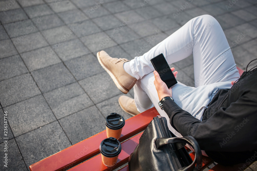 Obraz premium Cut view of man in jeans sitting on bench outside and hold black phone. There re two cups and leather bag.