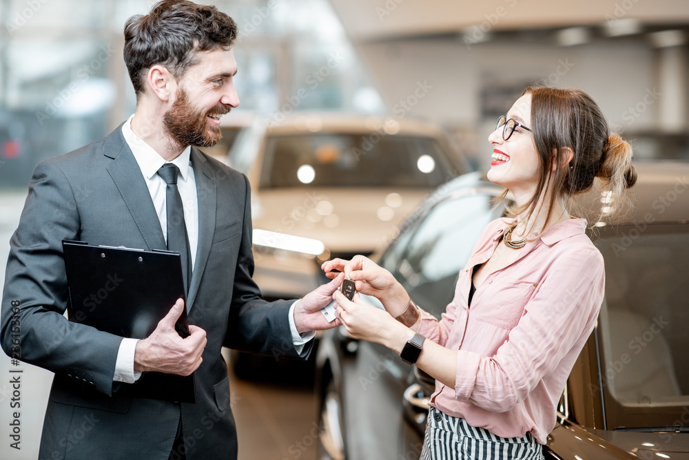 Fototapeta premium Salesperson giving keys to a young happy woman in the showroom with luxury cars