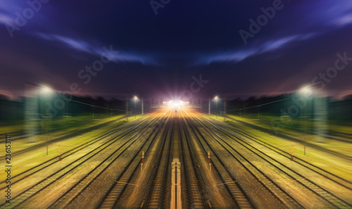 Bahnschienen mit symetrischen Linien - Bahnhof mit Lichter und Zug in der Ferne (Nachtaufnahme - Blaue Stunde)