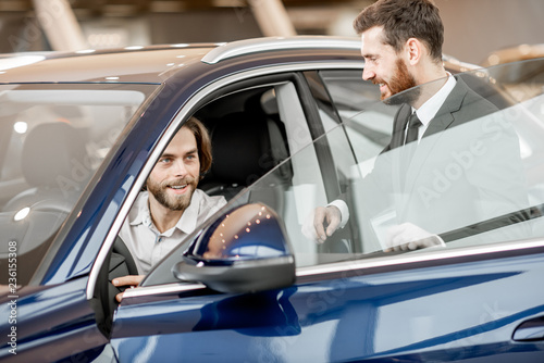 Salesperson helping young man to choose a new car sitting on the driver seat of a luxury car in the showroom