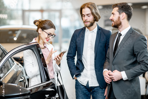 Salesperson standing with young business couple near the luxury car in the showroom
