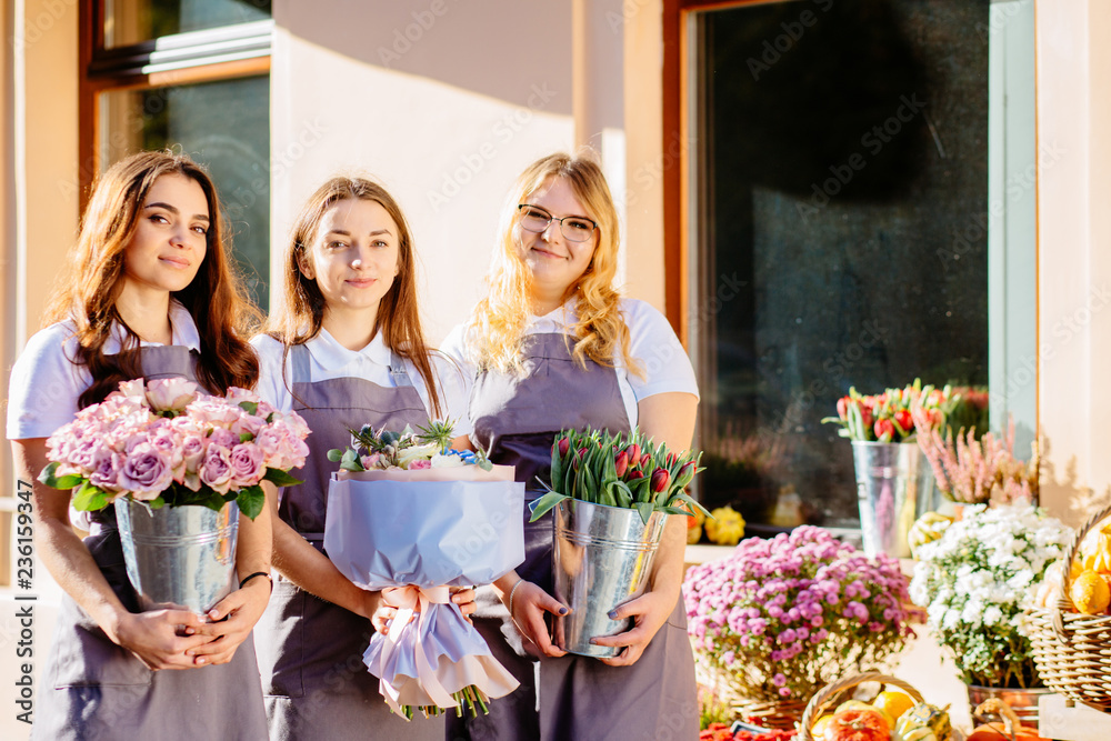 POrtrait of three friendly florists female team holding different flowers in pots outdoor near shop window with autumn decoration of their floral shop. Small business concept.