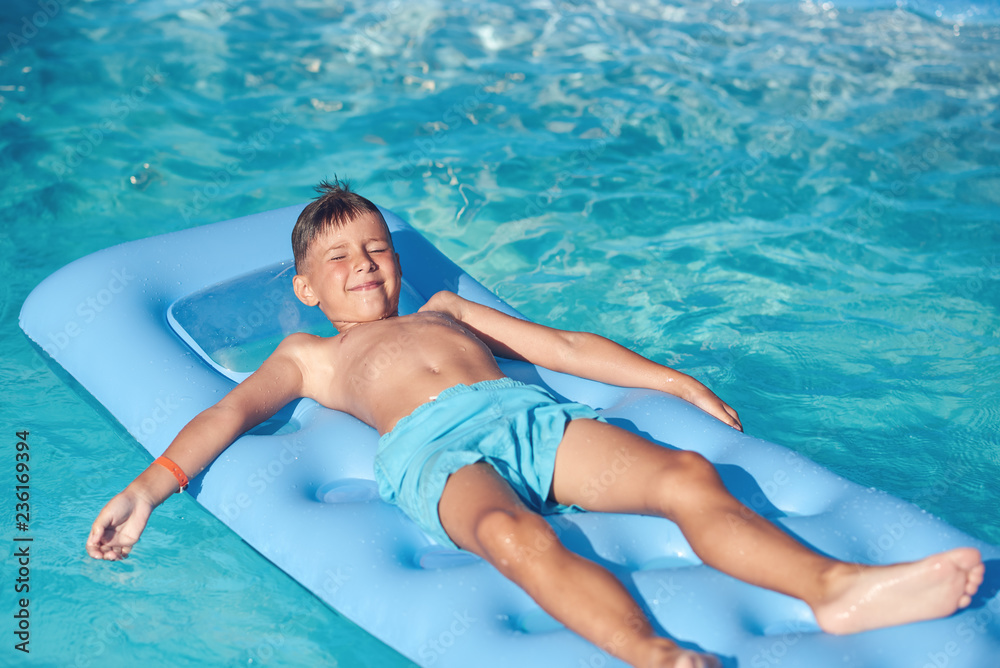 Smiling Caucasian boy with closed eyes laying on inflatable mattress at hotel swimming pool.