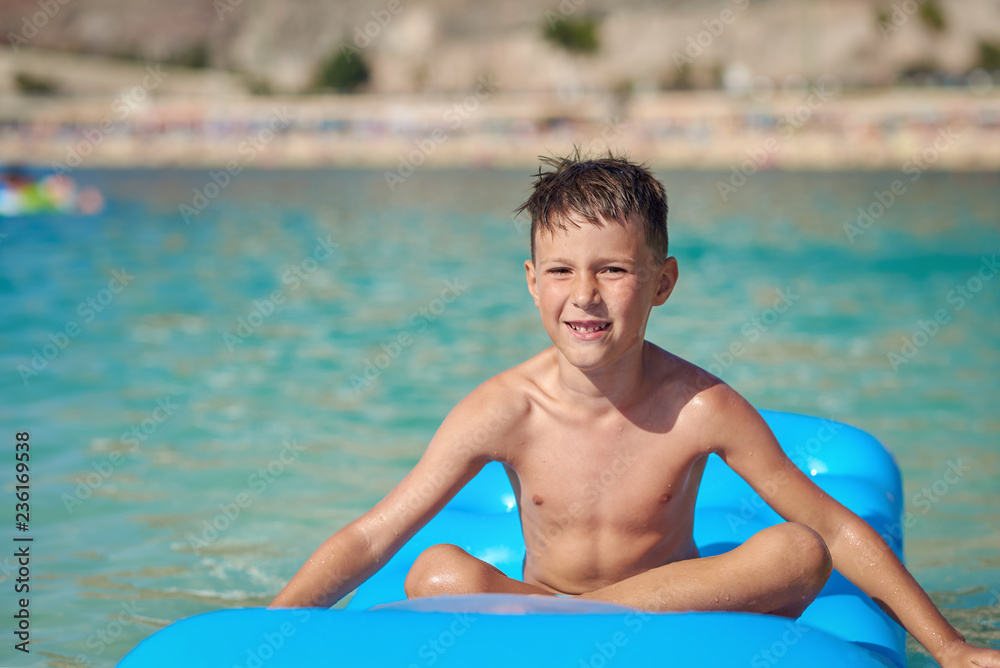 Cute European boy is sitting and swimming on the blue air matress, he is enjoying his holidays and smiling to the camera.