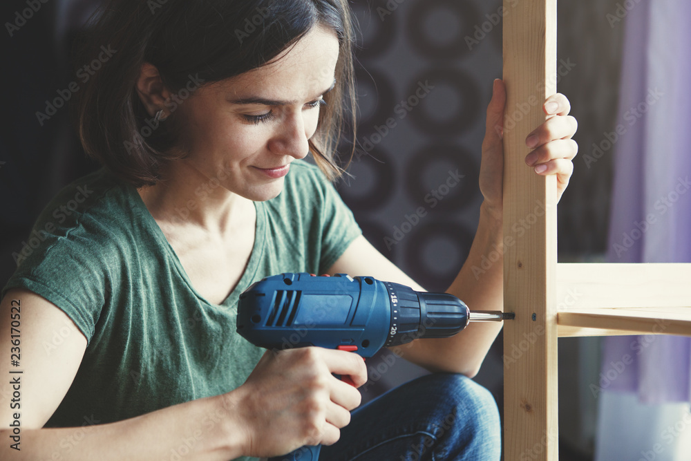 beautiful young woman holding screwdriver and repairing or making wooden shelf - obrazy, fototapety, plakaty 