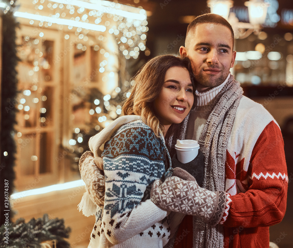 A young romantic couple wearing warm clothes outdoor at Christmas time, hugging and warming with hot coffee in evening street decorated with beautiful lights.