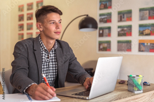 Portrait of the thinking young man who is sitting at the table and working with laptop and writing in the notebook indoor © Myroslava