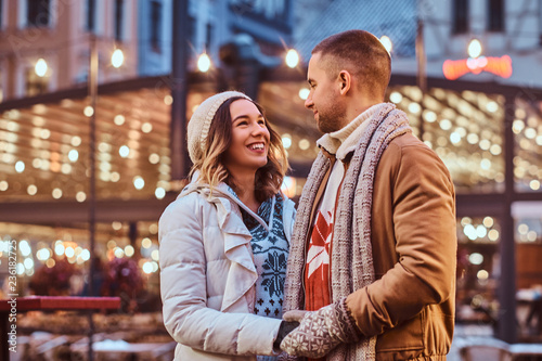 A young romantic couple holding hands and looking at each other while standing on the street at Christmas time, enjoying spending time together.