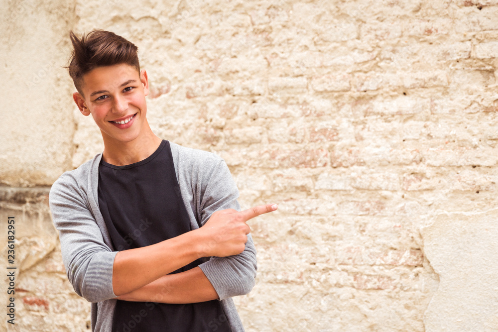Portrait of the smiling young man who is standing outdoor near the old house wall and showing aside by forefinger Portrait of the smiling young man who is standing outdoor near the old house wall and showing aside by forefinger