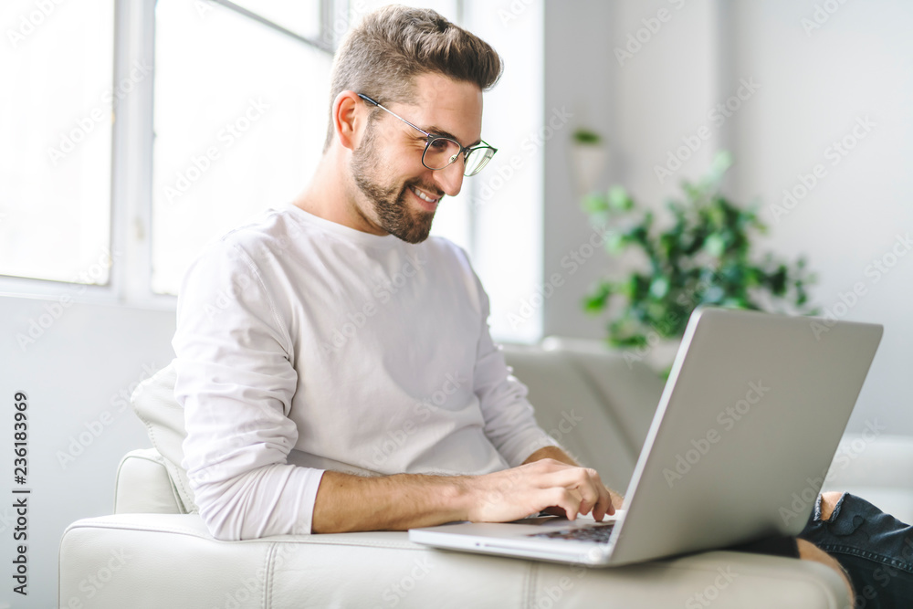 A Young attractive guy is browsing at his laptop, sitting at home on the cozy beige sofa at home, wearing casual outfit A Young attractive guy is browsing at his laptop, sitting at home on the cozy beige sofa at home, wearing casual outfit