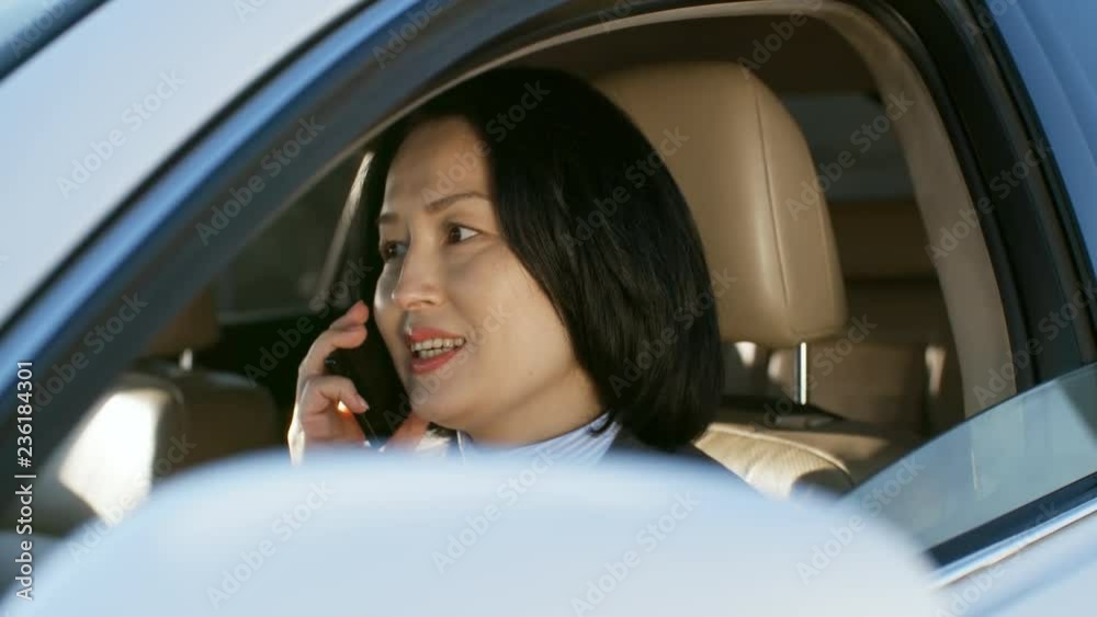 Medium shot of middle-aged Asian businesswoman having phone conversation while sitting in car Medium shot of middle-aged Asian businesswoman having phone conversation while sitting in car