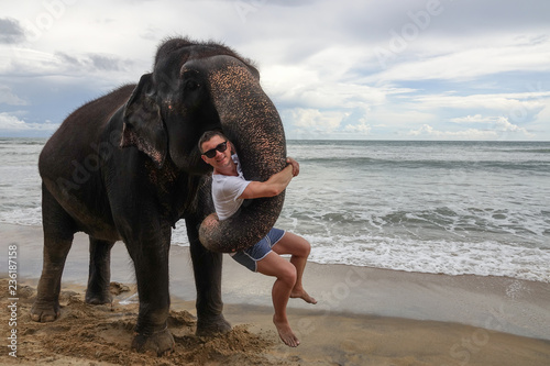 Portrait of a young guy with an elephant on the background of a tropical ocean beach. Elephant hugs a young man with his trunk. Tropical coast of Sri Lanka.