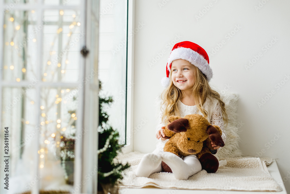 Little girl in the hat of Santa Claus with a plush deer sitting on the window. A child looks out the window and is waiting for Christmas, Santa Claus Little girl in the hat of Santa Claus with a plush deer sitting on the window. A child looks out the window and is waiting for Christmas, Santa Claus