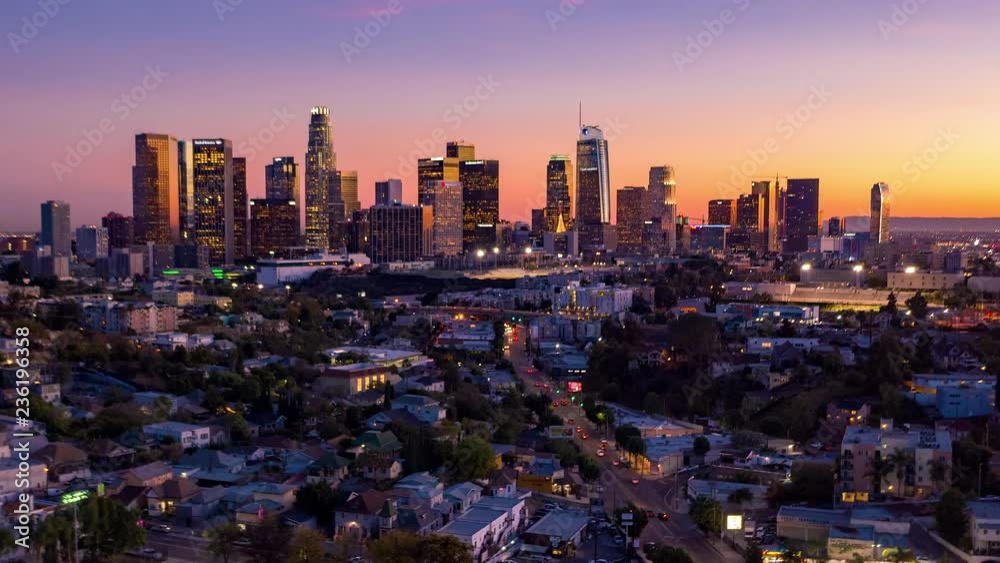 Aerial time lapse of downtown Los Angeles skyline with tall buildings and skyscrapers and freeway traffic below at twilight.