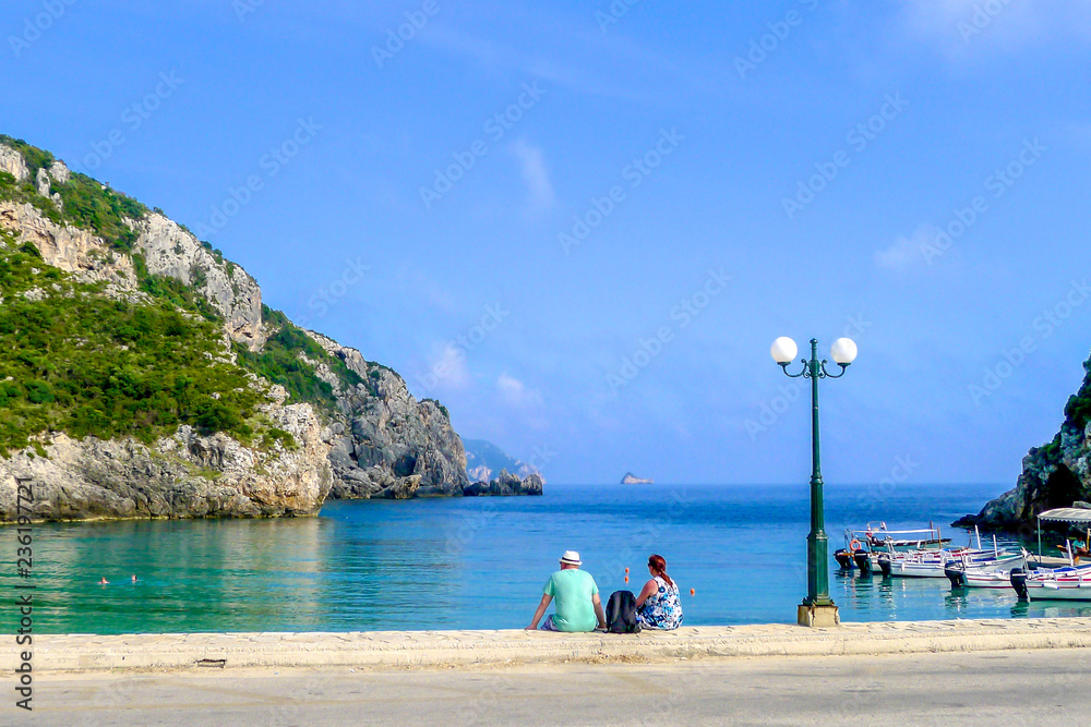 A couple of tourists sit on the waterfront overlooking the turquoise blue waters of Paleokastritsa beach in on the Greek island of Corfu, also known as Kerkyra, in the Ionian sea