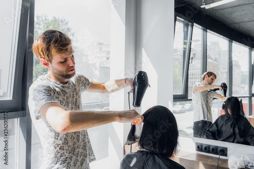 Young woman in a hair salon. Handsome male hairdresser drying hair with hair dryer