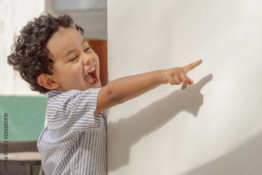 A curly hair little boy looks at a distance behind a white wall with a big  laugh pointing his finger. Stock Photo | Adobe Stock
