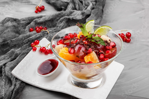 Fruit salad with banana, orange, grapes and pomegranate in glass bowl on grey background. Healthy breakfast