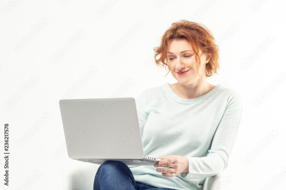 Attractive red haired mature smiling woman sitting in the chair with a laptop on her knees, working or watching something against white background Attractive red haired mature smiling woman sitting in the chair with a laptop on her knees, working or watching something against white background