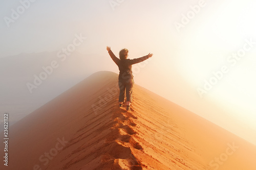 Person standing on top of dune in desert and looking at rising sun in mist with hands up, travel in Africa, Namibia 