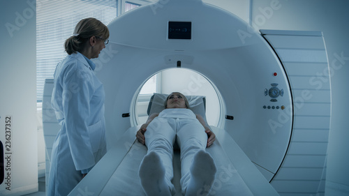 In Medical Laboratory Female Radiologist Controls MRI or CT or PET Scan with Female Patient Undergoing Procedure. Doctor Conducts Emergency Scanning with Advanced Medical Technologies. In Blue Tone.