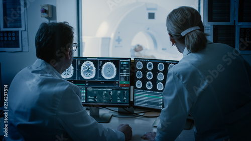 In Control Room Doctor and Radiologist Discuss Diagnosis while Watching Procedure and Monitors Showing Brain Scans Results, In the Background Patient Undergoes MRI or CT Scan Procedure.