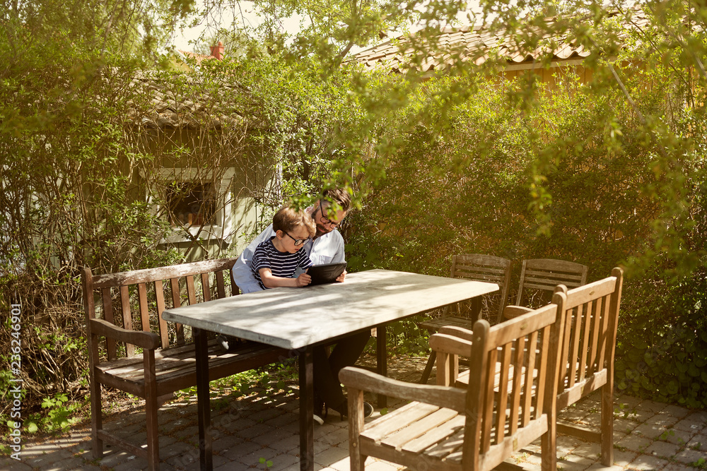 Mid adult man and boy use a device at an outdoor table in Sweden Mid adult man and boy use a device at an outdoor table in Sweden