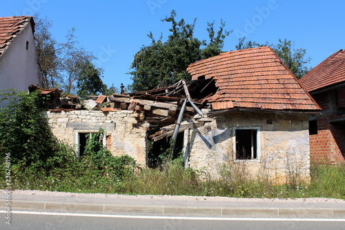 Small family brick house with destroyed roof, doors and windows during war completely overgrown with crawler plants and surrounded with high uncut grass next to paved road with high trees and clear bl
